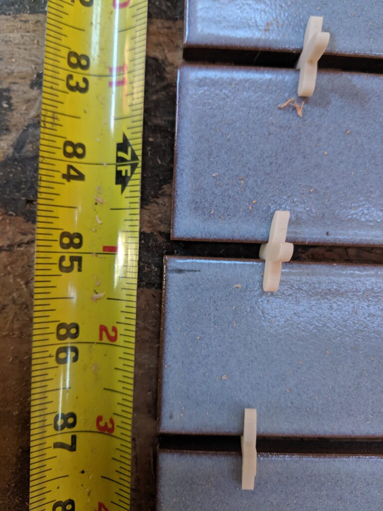 Close-up of handmade blue tiles with spacers next to a tape measure, the joints falling between round numbers